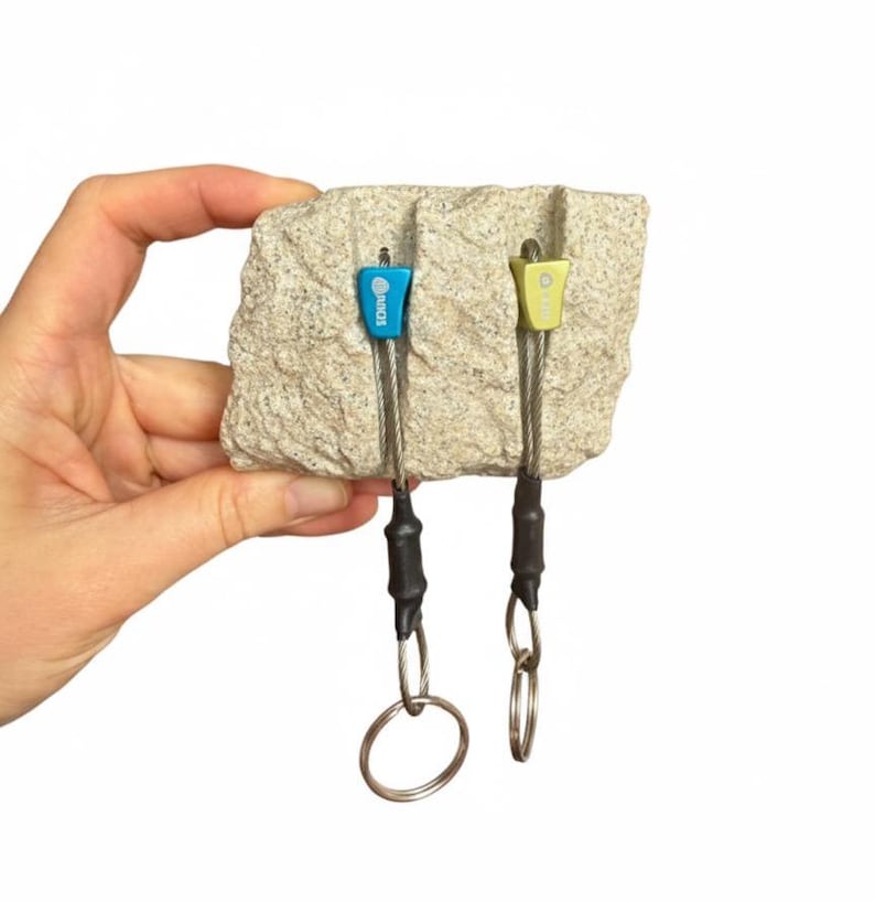 May include: A light blue and a light green climbing nut, each attached to a steel cable with a black heat-shrink sleeve and a metal ring, inserted into vertical grooves on a rectangular piece of textured, light beige granite rock.