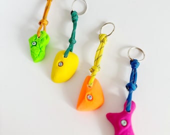 May include: Four colorful rock climbing holds attached to keychains. The holds are green, yellow, orange, and pink. Each hold has a small metal screw in the center.