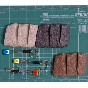 May include: Three rectangular stone-like blocks with three vertical grooves each, in beige, dark grey, and brown. Three metal cable keychains with coloured aluminium ends in blue, yellow, and orange, each featuring a circular steel ring.