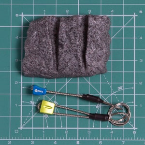 May include: A dark grey, textured rock with three vertical grooves positioned above two climbing nuts. The blue nut is labelled NIKIS, and the yellow nut is attached to a steel cable loop. The objects are placed on a green cutting mat with a grid.