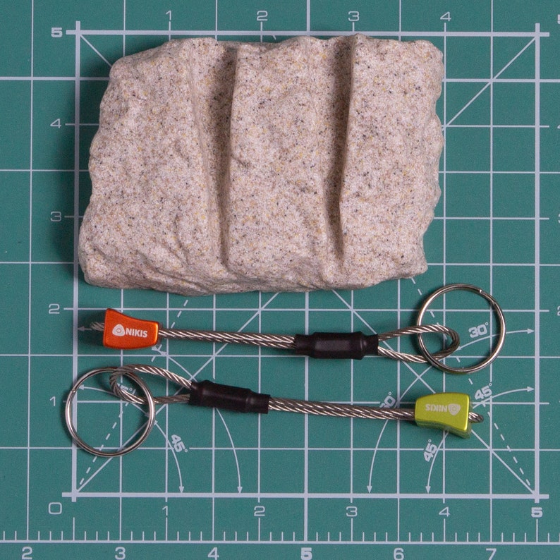 May include: A beige textured rock with two vertical grooves positioned above two metal keychains. Each keychain features a braided steel cable, a black heat-shrink sleeve, a silver split ring, and a small orange or lime green NIKIS branded component.