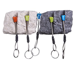 May include: Four climbing nuts with coloured metal heads in orange, lime green, and blue, each attached to a flexible steel cable and a circular metal ring. The nuts are positioned against two textured rock-like blocks, one beige and one dark grey.