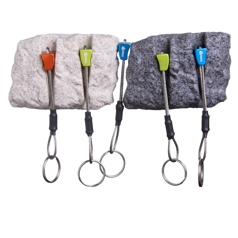 May include: Four climbing nuts with coloured metal heads in orange, lime green, and blue, each attached to a flexible steel cable and a circular metal ring. The nuts are positioned against two textured rock-like blocks, one beige and one dark grey.