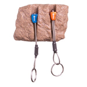 May include: Two climbing nuts, one blue and one orange, each labelled NIKIS, are inserted into vertical cracks in a textured brown rock-like block. Each nut features a flexible steel cable stem ending in a circular metal ring for attachment.