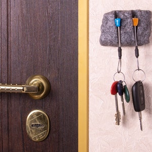 May include: A dark brown wooden door with a gold-toned metal handle and keyhole plate, positioned next to a grey stone-textured wall-mounted key holder. The holder features two blue and orange plastic pegs holding metal key rings with various keys.