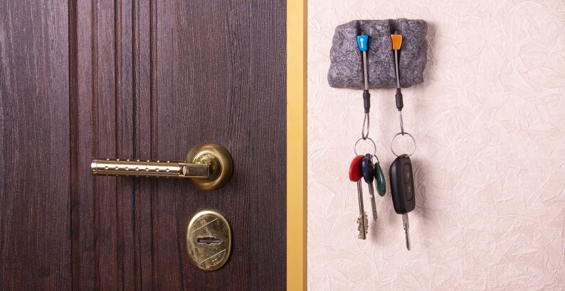 May include: A dark brown wooden door with a gold-toned metal handle and keyhole plate, positioned next to a grey stone-textured wall-mounted key holder. The holder features two blue and orange plastic pegs holding metal key rings with various keys.