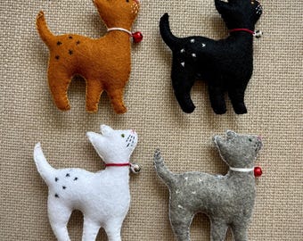 May include: Four felt cat ornaments in shades of brown, black, white, and gray. Each cat has embroidered details, a small bell collar, and star accents. The ornaments are arranged on a textured, neutral-colored surface.