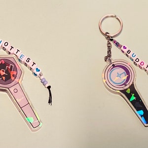 May include: Two iridescent keychains with beaded charms. The keychain on the left features a black and white light stick with a hexagonal head and the word "HOTTEST" spelled out in white beads. The keychain on the right features a black and white light stick with a round head and the word "BUDDY" spelled out in white beads.