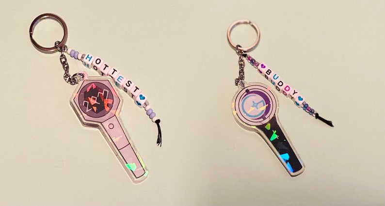May include: Two iridescent keychains with beaded charms. The keychain on the left features a black and white light stick with a hexagonal head and the word "HOTTEST" spelled out in white beads. The keychain on the right features a black and white light stick with a round head and the word "BUDDY" spelled out in white beads.
