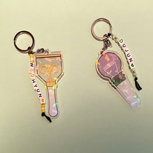 May include: Two iridescent keychains with beaded charms. One keychain features a lightbulb with the word "HIGHLIGHT" and the other features a microphone with the infinity symbol. The charms spell out "DUJUN" and "WOO HYUN" respectively.