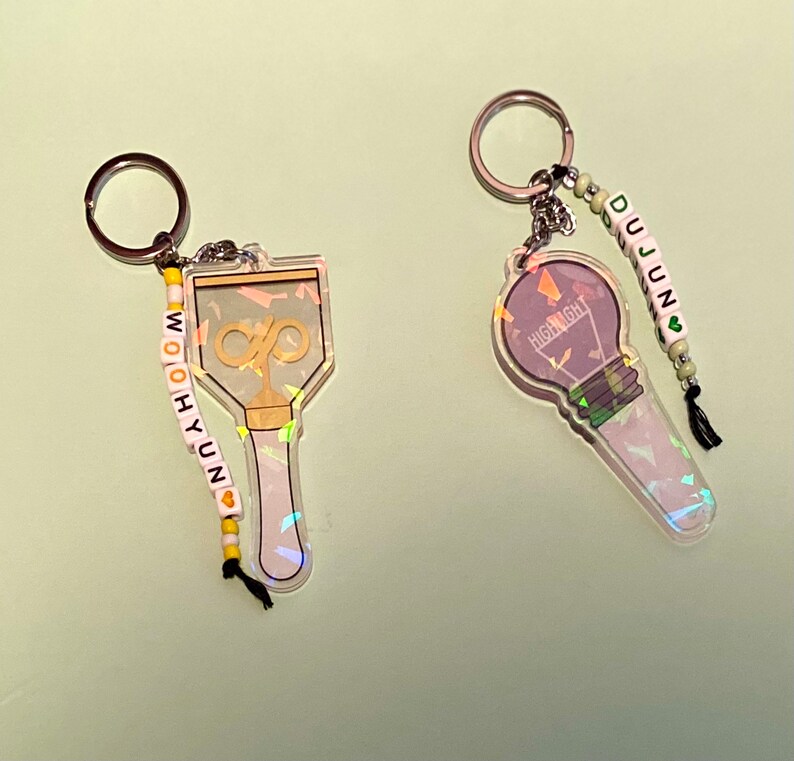 May include: Two iridescent keychains with beaded charms. One keychain features a lightbulb with the word "HIGHLIGHT" and the other features a microphone with the infinity symbol. The charms spell out "DUJUN" and "WOO HYUN" respectively.