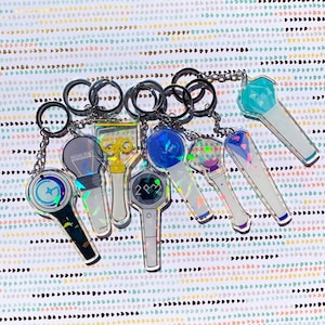 May include: A set of eight iridescent keychains featuring different K-pop group logos. The keychains are made of clear acrylic and have a holographic finish. The logos include "Highlight", "2PM", and a blue and white star.
