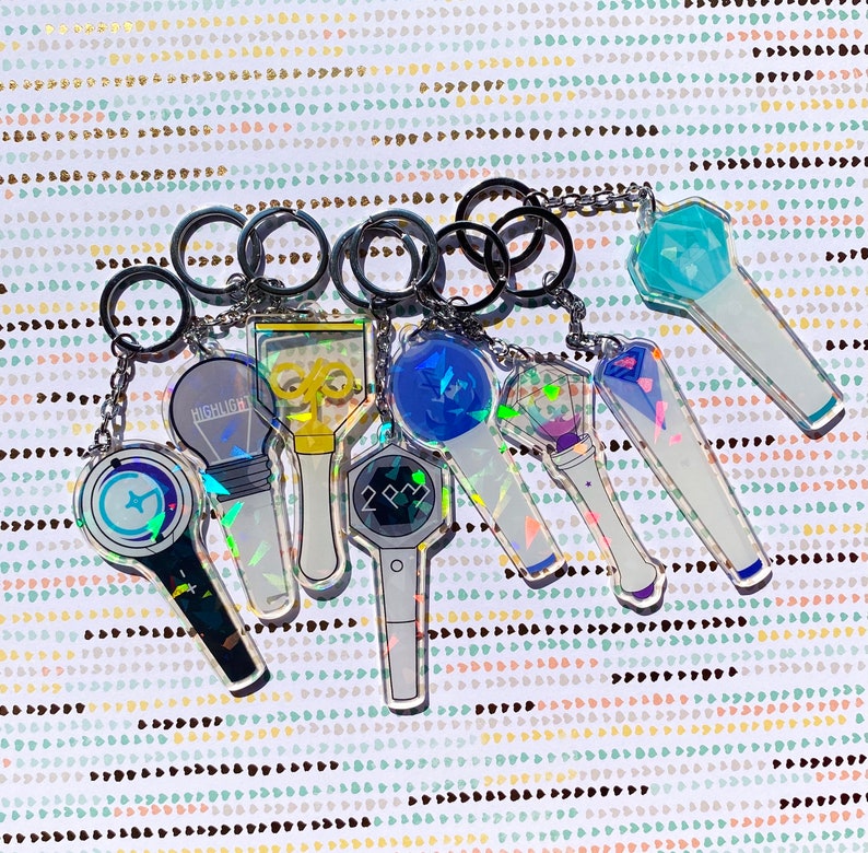 May include: A set of eight iridescent keychains featuring different K-pop group logos. The keychains are made of clear acrylic and have a holographic finish. The logos include "Highlight", "2PM", and a blue and white star.
