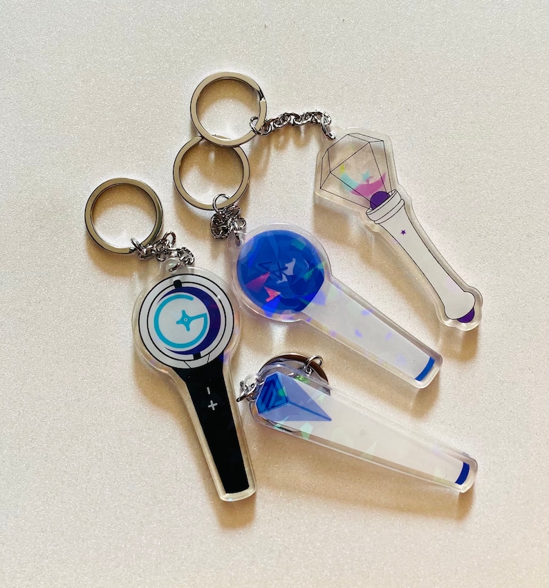 May include: Four different light-up keychain designs. The keychains are made of clear acrylic with a holographic finish. The designs include a blue and white star, a blue and white circle with a star, a blue and white diamond, and a blue and white circle with a star. The keychains are attached to silver key rings.