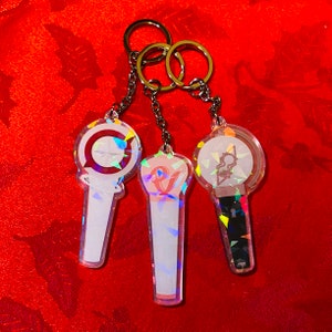 May include: Three iridescent keychains with different designs. Each keychain features a clear plastic charm with a holographic finish. The charms are shaped like light-up concert sticks and have different colored designs.