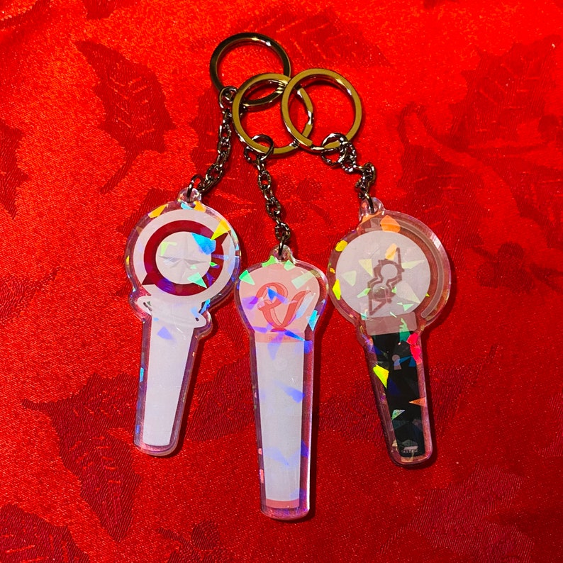 May include: Three iridescent keychains with different designs. Each keychain features a clear plastic charm with a holographic finish. The charms are shaped like light-up concert sticks and have different colored designs.