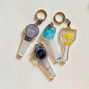 May include: Four different light stick keychains. The first is a clear light stick with a light bulb design and the word "HIGHLIGHT" printed on it. The second is a clear light stick with a blue diamond design. The third is a clear light stick with a yellow star design. The fourth is a clear light stick with a black and white design and the letters "2PM" printed on it.