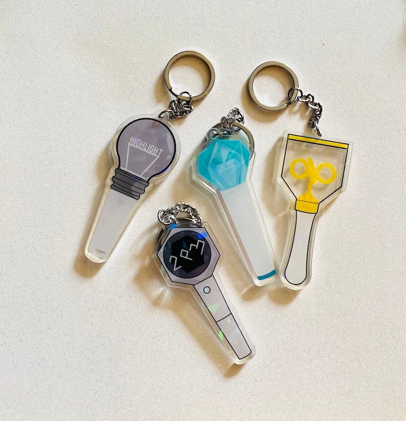 May include: Four different light stick keychains. The first is a clear light stick with a light bulb design and the word "HIGHLIGHT" printed on it. The second is a clear light stick with a blue diamond design. The third is a clear light stick with a yellow star design. The fourth is a clear light stick with a black and white design and the letters "2PM" printed on it.