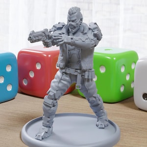 Peut inclure: Une figurine d'action futuriste en plastique gris imprimée en 3D tenant une arme à feu. La figurine est posée sur une base grise et est entourée de dés colorés.