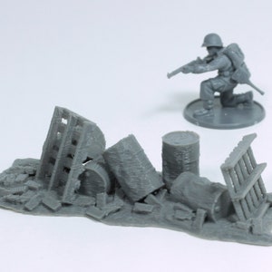 Puede incluir: Una figura de soldado en miniatura de plástico gris que sostiene un rifle, de pie cerca de una pila de escombros de plástico gris, incluidos barriles, una valla y otros escombros.