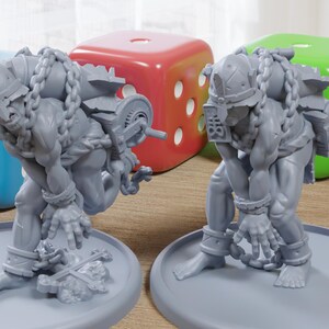 May include: Two gray, detailed figurines of muscular figures with chains and helmets, posed on gray circular bases. Large, colorful dice in red, green, and blue are in the background. The figurines appear to be fantasy or gaming related.