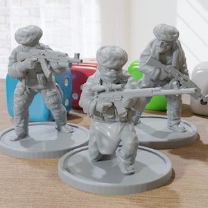 May include: Three gray miniature figurines of soldiers in combat gear, each holding a rifle. The figurines are on circular bases, with dice in the background. The soldiers are wearing head coverings and tactical vests.