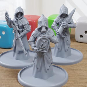 May include: Three gray miniature figures of hooded characters, each on a circular base, holding weapons or objects. The figures are surrounded by colorful dice: blue, red, green, and white. The scene is set on a wooden surface.