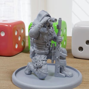 Puede incluir: Una figura en miniatura de plástico gris de una persona que lleva una chaqueta con capucha y sostiene un rifle. La figura está arrodillada sobre una base redonda.