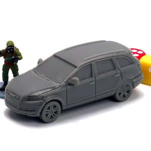 May include: A gray toy car model of a SUV with a person in a gas mask standing next to it. The car is on a white surface with three dice next to it.