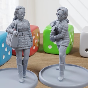 May include: Two grey figurines of women wearing sunglasses, coats, and boots, each holding a bag. They stand on grey circular bases, with large colourful dice in the background. The dice are blue, red, green, and white.