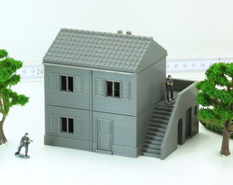 May include: A gray two-story model building with a tiled roof, windows, and a door. The building has a set of stairs leading up to a balcony. There are two small figures standing on the balcony and a small green tree in the background.
