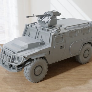 May include: A gray military armored vehicle with a machine gun mounted on the roof. The vehicle has a large front bumper with a grill and headlights. The vehicle is sitting on a wooden surface.