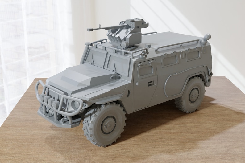 May include: A gray military armored vehicle with a machine gun mounted on the roof. The vehicle has a large front bumper with a grill and headlights. The vehicle is sitting on a wooden surface.