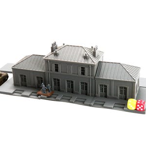 Op de afbeelding: Een grijs model van een treinstation met een tank en miniatuurfiguren. Het station heeft meerdere ramen en deuren. Twee dobbelstenen, één geel en één rood, liggen op het perron.
