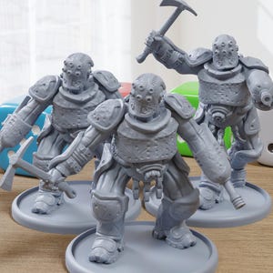 May include: Three gray, futuristic warrior figurines with detailed armor and helmets, each holding a weapon. The figures stand on circular bases, with dice in the background. The scene is set on a wooden surface.