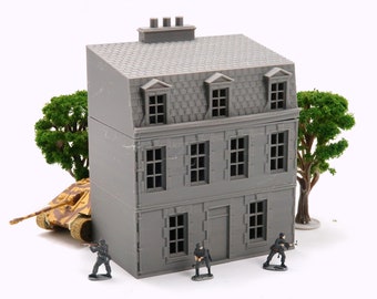 May include: A gray miniature building with a sloped roof, dormer windows, and a chimney. A tan and brown toy tank is positioned near the building, with miniature soldiers nearby. Green trees are also present.