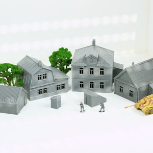 Op de afbeelding: Een model dorpset met grijze gebouwen, bomen en een kleine tank. De gebouwen zijn gemaakt van plastic en hebben een realistische uitstraling. De set bevat een verscheidenheid aan gebouwen, waaronder huizen, een schuur en een buitentoilet. De tank is lichtgeel en heeft een camouflagepatroon.