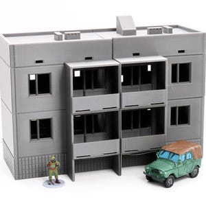 Op de afbeelding: Een grijs, drielaags gebouw met balkons en ramen. Een groene speelgoedauto staat voor het gebouw. Een klein groen figuurtje staat voor het gebouw.