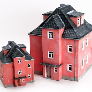 May include: Two miniature red houses with black roofs and white window frames. The larger house has a multi-level design with a chimney. The smaller house has a simpler, two-storey structure. Both are set against a white background.