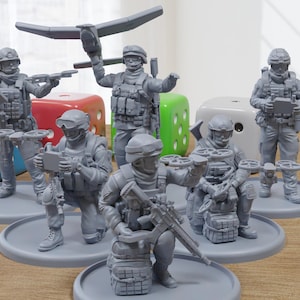 May include: Six gray plastic figurines of soldiers wearing camouflage gear and holding weapons. The soldiers are standing on gray circular bases. The bases are on a wooden surface.