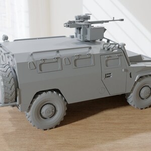 May include: A gray military armored vehicle with a mounted machine gun on top. The vehicle has a spare tire on the back and a roll cage.