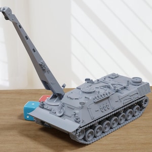 以下が含まれることがあります： クレーンアームを備えた軍用車両の灰色の模型。車両には詳細なトラックと複雑な上部構造があります。赤と青の立方体が車両の前にあります。模型は木製の表面にあります。