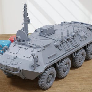 Op de afbeelding: Een grijs model van een militaire gepantserde transportwagen met een opgeheven antenne en een grote schotel aan de zijkant. Het voertuig heeft acht wielen en staat op een houten oppervlak.