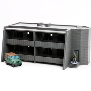 Op de afbeelding: Een grijs, twee verdiepingen tellend gebouwmodel met ramen en een deur. Het gebouw heeft een plat dak en een getextureerde buitenkant. Een kleine groene speelgoedauto staat voor het gebouw.