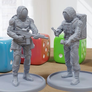 Può includere: Due figurine grigie di plastica di astronauti, una con una pistola e l'altra con uno strumento, in piedi su basi grigie. Le figurine sono su un tavolo di legno con quattro dadi sullo sfondo.