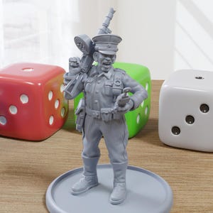 May include: A gray plastic miniature figurine of a soldier wearing a hat and holding a gun. The soldier is standing on a gray base. There are three dice in the background, one red, one green, and one gray.