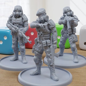 May include: Three gray plastic miniature figures of soldiers wearing gas masks and holding rifles. The figures are standing on gray circular bases. The figures are on a wooden surface with three dice in the background.