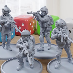 May include: Four gray miniature soldier figurines, each posed with a rifle, stand on individual gray circular bases. Colorful dice in blue, red, and green are in the background, along with a white die. The figurines are detailed with tactical gear.
