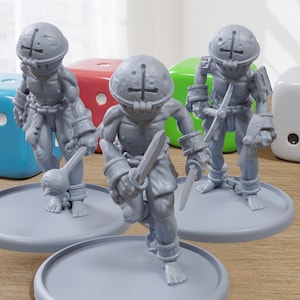 May include: Three gray miniature figures of armored characters with weapons, standing on gray circular bases. Colorful dice in blue, red, green, and white are in the background. The figures have helmets and are posed as if in action.