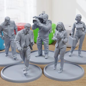 Puede incluir: Seis figuritas grises pintadas de personas, cada una de pie sobre una base circular gris. Las figuritas representan a un grupo de reporteros, incluyendo un camarógrafo, una mujer sosteniendo un micrófono y un hombre sosteniendo un maletín.